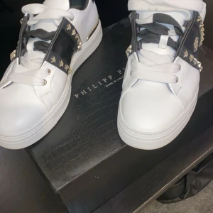 Philipp Plein vita sneakers med nitar - Snygga vita sneakers från Philipp Plein med svarta detaljer och guldfärgade nitar. Skorna har bred vit sula, snörning och svart läderdetalj med logga på sidan. Guldiga accenter vid hälen och coola stjärnnitar ger en lyxig vibe.