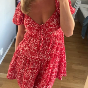 Röd blommig kortklänning med knyt - Supersöt röd kortklänning med vita blommor, v-ringning och knyt framtill. Puffiga korta ärmar och volangig kjol ger en romantisk vibe. Perfekt för varma dagar och sommarhäng.