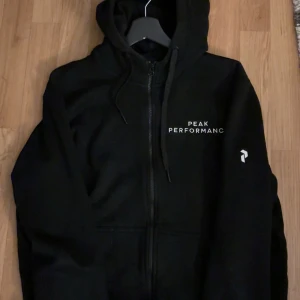Svart hoodie från Peak Performance - Svart hoodie från Peak Performance med dragkedja framtill och vit logga på bröstet. Tröjan har huva med snörning och fickor på sidorna. Perfekt för en avslappnad och sportig stil.