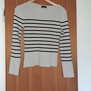 Randig långärmad tröja från Kookai - Supersnygg ribbad tröja från Kookai i ljusgrått med svarta horisontella ränder. Långärmad och figurnära passform, perfekt till jeans eller kjol. Mjuk och stretchig, med klassisk rund halsringning. Enkel men ändå trendig stil.