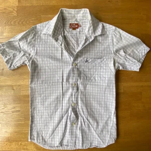 Vit rutig skjorta från Encore, M - Vit skjorta från Encore i storlek M med  rutmönster, klassisk krage och korta ärmar. Slim fit-modell med knappstängning framtill och bröstficka med broderad logga. Perfekt för dig som gillar stilrena och fräscha skjortor.