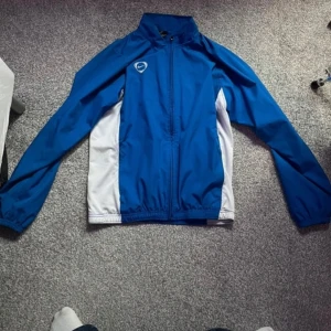 Blå Nike vindjacka med vita detaljer - Snygg blå vintage vindjacka från Nike med vita paneler längs sidorna och logga på bröstet. Jackan har hel dragkedja och meshfoder på insidan för extra komfort. Perfekt för dig som gillar sportig stil och vill ha något lätt och funktionellt och streety, diskuterar gärna priset, passar S.