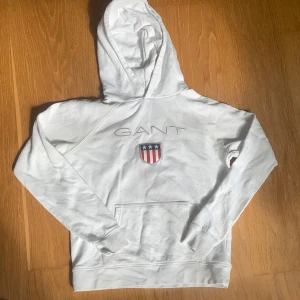 Vit hoodie från GANT med logga - Vit hoodie från GANT med broderad logga och amerikanskinspirerat emblem på bröstet. Tröjan har huva, lång ärm och en stor magficka framtill. Tillverkad i mjuk bomull, perfekt för chill dagar. Passar dig som är 13-14 år och gillar klassisk stil.