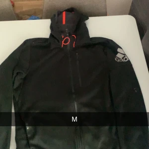 Svart vindjacka från Adidas - Säljer en svart vindjacka från Adidas med huva och dragkedja framtill. Jackan har röda detaljer på huvan och snörning, samt Adidas-logga på ena ärmen. Perfekt för dig som gillar sportig stil och vill ha något snyggt och funktionellt.