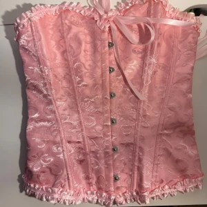 Rosa korsett med spets och snörning - Supersöt rosa korsett med glansigt tyg och broderade detaljer. Den har snörning i ryggen, knappar framtill och volangkant upptill och nedtill. Satinband och romantisk spets ger en extra girly vibe. Perfekt för att skapa en statement look! Jag använde den för att vara Cupid på Halloween.     Size S/36. ( använd en gång)