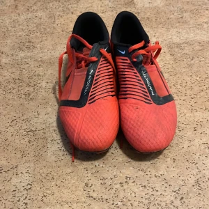 Nike Phantom VNM röda fotbollsskor - Säljer ett par Nike Phantom VNM fotbollsskor i en riktigt snygg röd färg med svarta detaljer och snörning. Skorna har en rund tå och är designade för grym kontroll på planen. Ovandelen är i syntetmaterial och insidan är svart för extra kontrast. Perfekt för dig som vill sticka ut på matchen!