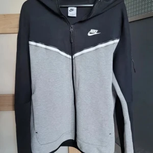 Nike tech-fleece svart, grå. - Nike hoodie i svart och ljusgrått med vit logga på bröstet. Tröjan har huva, hel dragkedja och långärmade ärmar. Snyggt designad med vit linje över bröstet och praktiska fickor. Perfekt för chill eller träning.