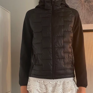 Svart pufferjacka med huva - Svart pufferjacka med quiltad framsida och rygg, släta ärmar och huva. Jackan har dragkedja framtill och en clean, sportig look. Perfekt för dig som gillar stilren design och vill ha en jacka som funkar till många olika outfits.