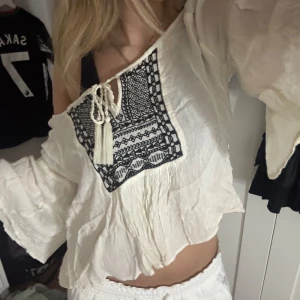 Vit blus - Säljer en luftig vit blus med svart broderat mönster framtill och snörning med tofsar vid halsen. Blusen har vida trekvartsärmar och är tillverkad i mjukt, tunt material som ger en chill vibe. Jättefin som overshirt till stranden, eller bara en mysig sommardag. Den behöver strykas.