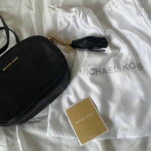 Svart axelväska från Michael Kors - Snygg svart axelväska från Michael Kors i skinn med guldfärgade kedjedetaljer och en dekorativ tofs. Väskan har oval form, dragkedja upptill och logga i guld på framsidan