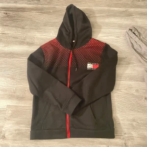 Svart hoodie från Tommy hill med röd dragkedja - Svart hoodie från Tommy hill med röd dragkedja och coolt prickigt mönster i rött över axlarna. Har huva med snörning och fickor framtill. Logga på bröstet i rött, vitt och grått. Perfekt för dig som gillar streetstyle och vill sticka ut.