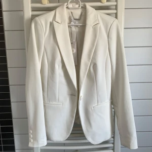 Vit kavaj med klassisk passform - Snygg vit kavaj med klassisk krage och en knapp framtill. Kavajen har figurnära passform, två fickor och dekorativa knappar vid ärmslutet. Materialet känns mjukt och har en slät yta, perfekt för en stilren look. Passar dig som gillar minimalistisk och fräsch stil.