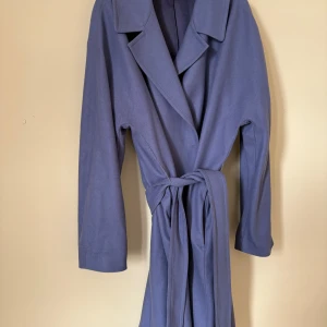 Blå kappa från ARKET med bälte - Snygg blå kappa från ARKET med klassisk krage och knytbälte i midjan. Långärmad och med stilren, rak passform. Materialet är mjukt och känns lyxigt, vävt i Italien av Fortex. Perfekt för dig som gillar minimalistisk och clean stil.