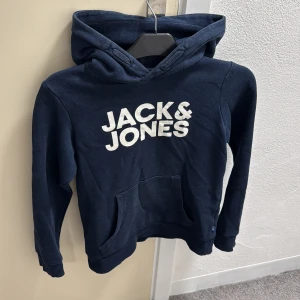 Mörkblå hoodie från Jack & Jones - Snygg mörkblå hoodie från Jack & Jones med stor vit logga på bröstet. Tröjan har huva, långärmade ärmar och en praktisk magficka framtill. Tillverkad i mjuk bomull som är skön att ha på sig. Perfekt för chill dagar och streetstyle.