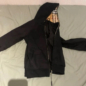 Svart hoodiejacka från Burberry - Svart hoodiejacka från Burberry med klassiskt rutig foder i huvan. Jackan har dragkedja, snörning i huvan och två fickor framtill. Perfekt för dig som gillar stilren design med en touch av lyx. Materialet är mjukt och bekvämt bomull. Men är liten och kanske inte som står M passar mer till S. Ingen ångresrätt eller retur.Oanvänd och helt ny skicka och fråga om ni är intresserade😁.