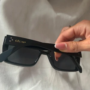 Svarta Céline solglasögon - Säljer ett par svarta solglasögon från Céline med breda skalmar och fyrkantig form. Glasögonen har en stilren design med mörka linser och diskret guldfärgad logga på sidan. Perfekta för dig som gillar en cool och trendig look.