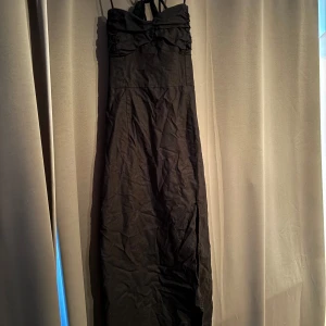 Svart långklänning från Zara - Svart långklänning från Zara med smala axelband och snygg knytning i ryggen. Klänningen har rynkade detaljer upptill och en smockad rygg för extra komfort. Materialet är mjukt och lätt, perfekt för dig som vill ha en stilren och elegant look. Har en slits som tyvärr spruckit lite upptill men inget som syns när den bärs. 