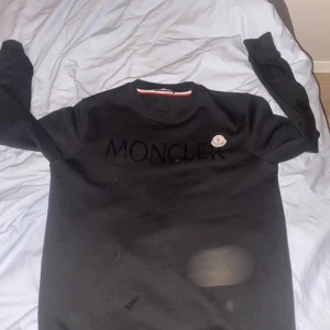 Svart Moncler sweatshirt  - Svart sweatshirt från Moncler med stor, svart Moncler-text på bröstet och klassisk logotyp-patch. Tröjan har rund hals, långa ärmar och är gjord i mjuk bomull. Perfekt för dig som gillar stilrena designer och exklusiva märken.