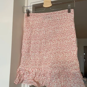 Rosa blommig kjol från Bik Bok - Supersöt kort kjol från Bik Bok i ljusrosa och vitt med blommigt mönster. Kjolen har smockad midja och volangkant nedtill