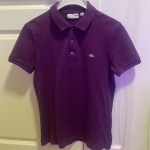 Lacoste piké - Slim fit, Snygg lila pikétröja från Lacoste i slim fit-modell. Klassisk krage, tre knappar framtill och den ikoniska krokodilloggan broderad på bröstet. Tillverkad i mjuk bomull som andas och har korta ärmar. Perfekt för dig som gillar stilren och sportig vibe.