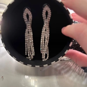 Glittriga silver örhängen med strass - Säljer ett par långa, glittriga örhängen i silverfärg med strass. De har en cool, hängande design med flera rader av stenar som ger extra bling och rörelse. Perfekta för att sticka ut och ge din look en lyxig touch.