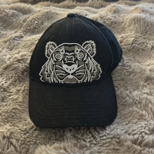 Svart Kenzo keps med tigerbroderi - Svart keps från Kenzo med ett stort, detaljerat tigerbroderi i silver och grått framtill. Klassisk böjd skärm och justerbar passform. Insidan har Kenzo-logga på banden och etikett. Perfekt för dig som vill sticka ut med en ikonisk streetstyle-accessoar.