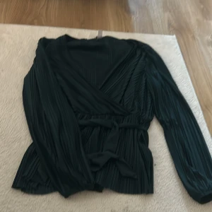 Mörkgrön omlottblus med knytband - Mörkgrön svart omlottblus med plisserade detaljer och knytband i midjan. Blusen har långärmade ärmar och ett elegant omlottliv som ger en schysst siluett. Perfekt för dig som gillar stilrena och trendiga plagg med lite extra edge.