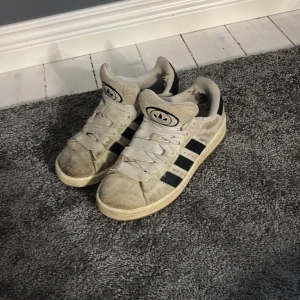 Adidas Campus sneakers i vitt och svart - Adidas Campus sneakers med klassiska svarta ränder på sidorna och vit ovandel. Skorna har rund tå, platt sula och snörning. Materialet är mestadels syntet och textil, med Adidas-logga på plösen. Det är hål innanför därav lågt pris. De kommer rengöras innan köp om man vill se hur de ser ut då får man skicka om bild.
