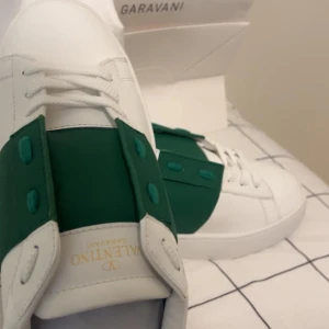 Valentino Garavani Open sneakers vit/grön - Snygga Valentino Garavani Open sneakers i vitt skinn med en bred grön läderpanel över snörningen. Klassisk rund tå och platt sula, med coola nitar på hälen. Perfekt för dig som vill sticka ut med stilrena och lyxiga sneakers.