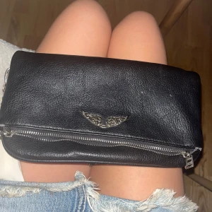 Äkta Zadig & Voltaire väska - Snygg svart axelväska i skinn från Zadig & Voltaire med silverfärgad kedja och dragkedjor. Väskan har ett coolt emblem med vingar framtill och flera fack med dragkedja både fram och bak. Perfekt storlek för det viktigaste och riktigt trendig look. I bilderna ser den platt ut men när man väll fyller den är den super cool!