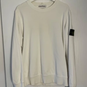 Vit sweatshirt från Stone Island - Vit långärmad sweatshirt från Stone Island med klassisk rund hals och ikonisk svart patch med gul och grön broderad logga på vänster ärm. Tröjan är i mjuk bomull och har ribbade muddar vid ärmslut och nederkant. Perfekt för en clean och stilren look.