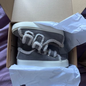 Grå sneakers från Inuikii med fårull storlek 39 - Unika grå sneakers från Inuikii med fluffiga vita detaljer i fårull och breda snörningar. Skorna har en platt vit sula och är tillverkade i mocka med mjuk insida. Perfekt för dig som vill ha en cool och mysig look under höst och vinter. Storlek 39, slutsålda på de flesta sidorna
