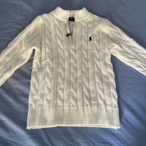 Vit stickad tröja Polo Ralph Lauren - Vit kabelstickad tröja från Polo Ralph Lauren med halv dragkedja och ribbad krage. Klassisk design med broderad logga på bröstet och snygga detaljer vid dragkedjan. Perfekt för dig som gillar stilrena och tidlösa plagg.