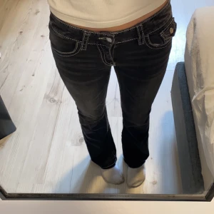 Svarta bootcut jeans med kontrastsömmar - Snygga svarta jeans med bootcut passform och tydliga vita kontrastsömmar. Modellen har låg midja, klassiska fickor fram och bak med lock och knappdetaljer. Jeansen är gjorda i jeansmaterial och har en cool, lite retro vibe. Använda typ 4 gånger💕