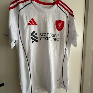 Liverpool FC Tröja - Snygg vit Liverpool FC fotbollströja från Adidas med rött klubbmärke och röda detaljer på axlarna. Tröjan har svart piping längs sidorna, korta ärmar och rund hals. Materialet är lätt och ventilerande polyester, perfekt för match eller träning.