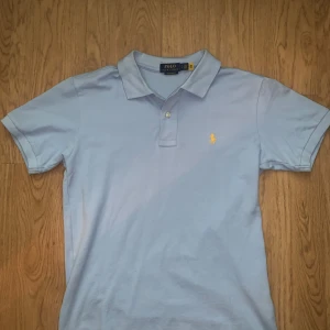 Ljusblå Polo Ralph Lauren t-shirt - Snygg ljusblå t-shirt från Polo Ralph Lauren i bomull med klassisk pikékrage och korta ärmar. Custom slim fit-modell med gul broderad logga på bröstet. Perfekt för dig som gillar stilrena och ikoniska plagg.