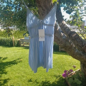 Ljusblå kortklänning från Zara - Supersöt ljusblå klänning från Zara i storlek S. Klänningen har tunna axelband, v-ringad front och är plisserad med smockdetaljer vid midjan och bysten. Perfekt för varma dagar och har en flowy, luftig känsla. Materialet är lätt och känns som polyester.