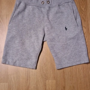 Grå sweatshorts från Polo Ralph Lauren - Grå mjuka sweatshorts från Polo Ralph Lauren med klassisk broderad logga på benet. Modellen har midwaist, snörning i midjan och två sidofickor. Perfekta för chill eller träning, tillverkade i bomull för skön komfort.