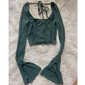 Grön croppad långärmad topp med knyt - Supersnygg mörkgrön croppad topp med långa trumpetärmar klockade ärmar och ribbad struktur. Toppen har en djup rundad halsringning med knyt i nacken och markerade sömmar framtill som ger en cool vibe. Perfekt för dig som gillar unika detaljer och trendig stil.