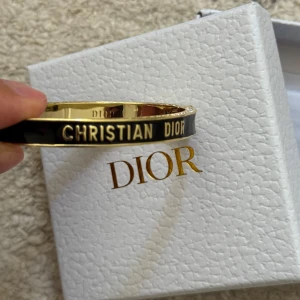 Svart och guld armband med strass  - Exklusivt armband i guldtonad metall med svart emalj och tydlig logotyp. Insidan är dekorerad med gnistrande stenar som ger extra lyxig känsla. Snyggt och stilrent smycke som verkligen sticker ut med sin eleganta design. Man öppnar det på sidan. Aldrig använt fel storlek för mig 