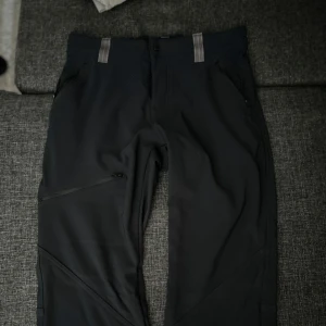Svarta täckbyxor från Columbia - Svarta täckbyxor från Columbia med gråa hällor och diskret logga på höften. Byxorna har dragkedja på benet och praktiska fickor. Materialet är slitstarkt och syntetiskt, perfekt för kalla dagar och utomhusaktiviteter. Snygg, rak passform som funkar till många stilar. Kostade 1000 men har ingen användning för dem.