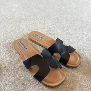 Svarta sandaler från Steve Madden - Snygga svarta sandaler från Steve Madden med breda remmar i skinn och öppen tå. Modellen har en platt sula och en stilren design med fyrkantig form framtill. Perfekta för varma dagar och enkel att matcha med olika outfits.