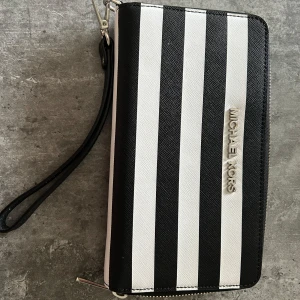 Randig plånbok från Michael Kors - Snygg och stor plånbok från Michael Kors med svartvita breda ränder och silverfärgad logotyp på framsidan. Plånboken har dragkedja runtom och en praktisk handledsrem. Insidan har flera fack för kort och sedlar. Perfekt för dig som gillar stilren design.