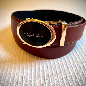 Brunt skärp från Ragole Lacas med gulddetaljer - Snyggt och brett brunt bälte från Ragole Lacas med exklusivt svart och guldigt spänne. Bältet är gjort i skinn och har dekorativa sömmar längs kanterna. Insidan är svart och spännet har en lyxig känsla med ovalt format och guldfärgade detaljer.