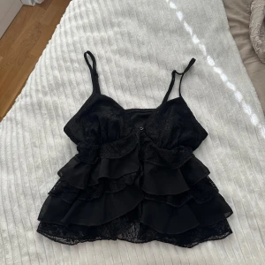 Svart volang topp med spets - Supersnygg svart topp med tunna axelband, flera lager volanger och detaljer i spets. Perfekt för dig som gillar en feminin och trendig look. Croppad modell som ger en cool vibe och passar till både jeans och kjol.