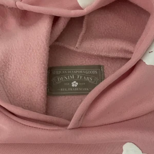 Rosa hoodie från Denim Tears med blommor - Säljer en rosa hoodie från Denim Tears med vita blommiga tryck över hela tröjan. Tröjan har huva, magficka och långa ärmar. Mjuk insida i fleece och loose passform. Perfekt för dig som gillar statement-plagg och coola detaljer.