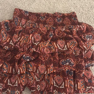 Mönstrad volangkjol från Lindex - Säljer en kort kjol från Lindex med flera volanger och ett färgstarkt paisleymönster i vinrött, orange, blått och vitt. Kjolen har bred resår i midjan och är gjord i ett lätt och luftigt material som passar perfekt till varma dagar. Passar till XS