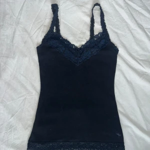 Marinblå spetslinne Abercrombie & Fitch - Supersnygg marinblå linnetopp från Abercrombie & Fitch med spetsdetaljer vid axelband, v-ringning och nederkant. Tunn bomullskvalitet, tight passform och broderad älglogga längst ner. Perfekt att styla med jeans eller kjol för en chill look. Tight men stretchiga hade passat S-L. 