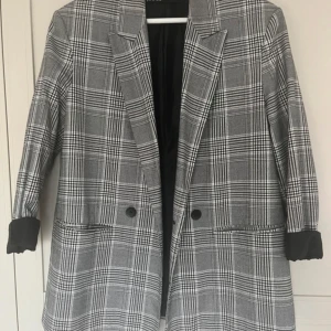 Rutig kavaj från Zara i grått - Snygg rutig kavaj från Zara i grå och vit ton med klassisk krage och två svarta knappar framtill. Kavajen har lång ärm och två fickor, perfekt för att lyfta din outfit med en trendig touch. Materialet är mjukt och känns bekvämt mot kroppen.