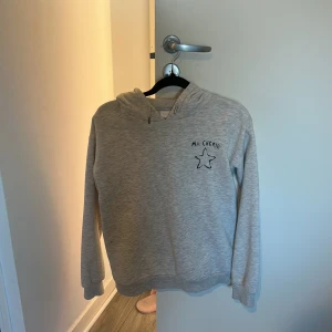 Grå hoodie med Ma Chérie-tryck - Säljer en ljusgrå hoodie med svart text 'Ma Chérie' och en stjärna tryckt på bröstet och ryggen. Tröjan har huva med snörning och långa ärmar. 